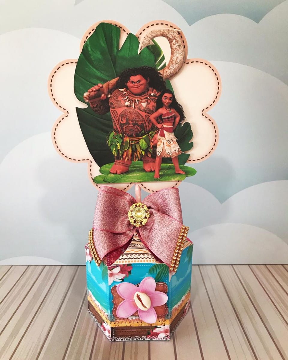 Arquivo Digital Centro de Mesa Moana Adulta PDF e Studio 1 Topo de bolo decorado com personagens de desenho animado, laço rosa brilhante e base estampada.