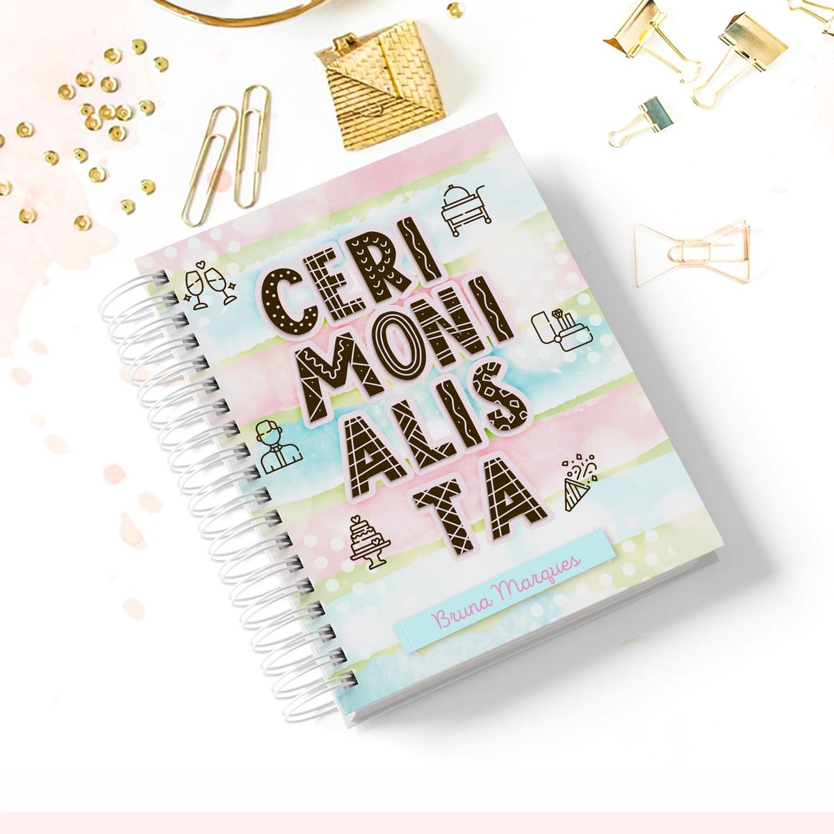 Caderno Digital Cerimonialista para Agendas Encantadoras 1 Caderno espiral colorido com capa decorada cercado por pequenos clipes dourados na mesa branca