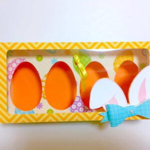 Caixa retangular amarela com visor para quatro ovos de Páscoa e orelhas decorativas.