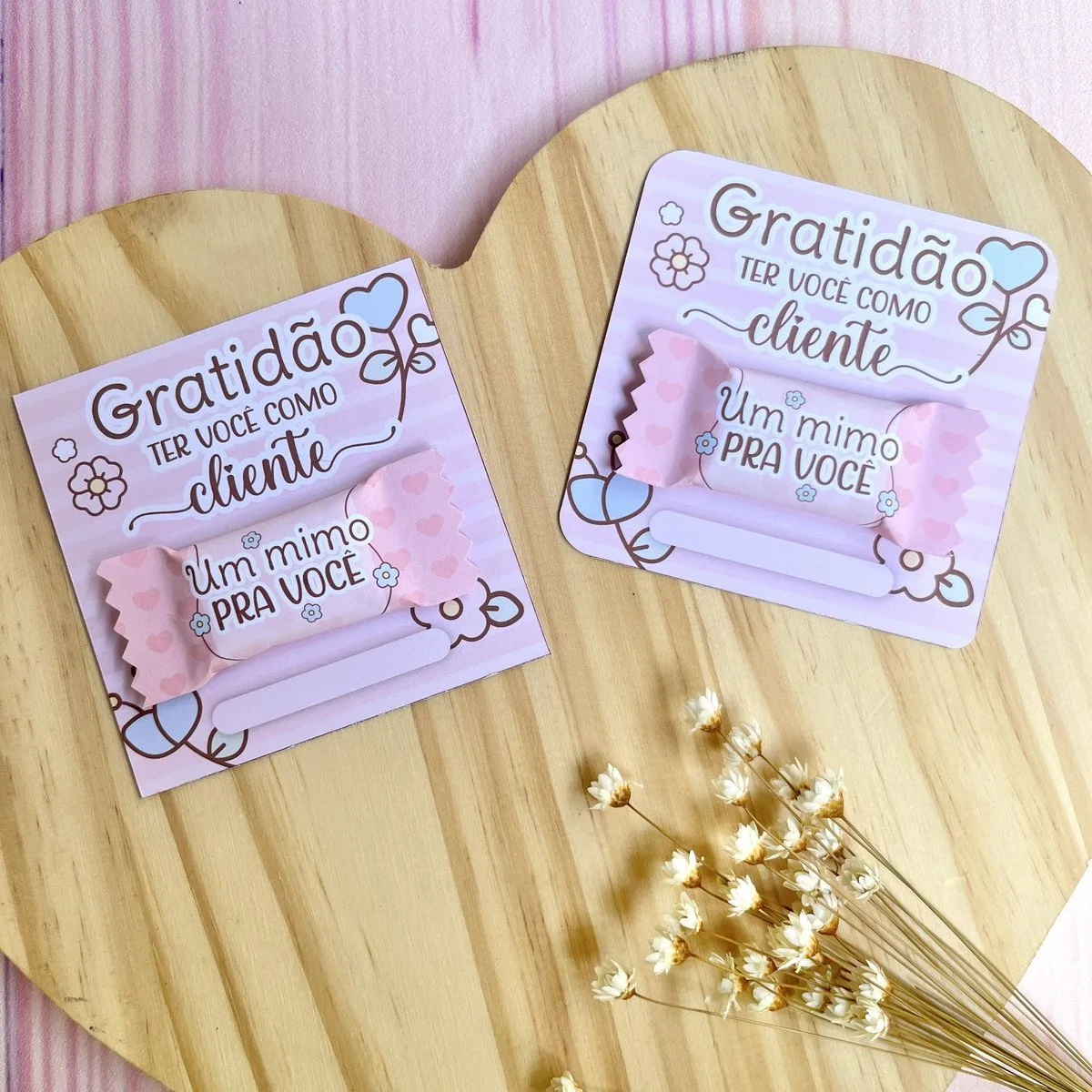 Card e Balinha Mimos Clientes 1 Dois cartões personalizados com doces embalados sobre base de madeira circular com flores secas.