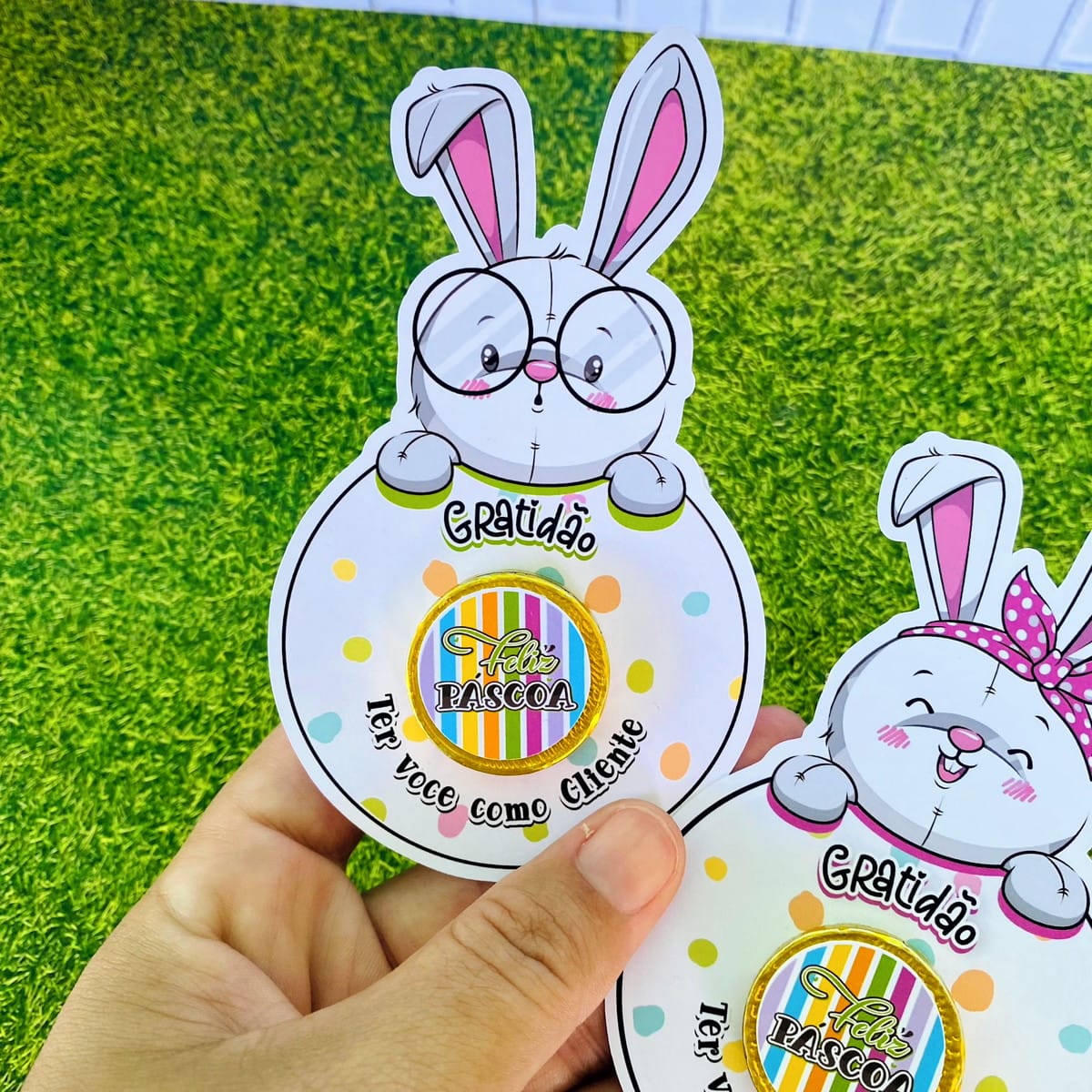 Card Moedinha de Páscoa Digital para Imprimir 4 Mão segurando dois cartões de papel com ilustrações de coelhos de Páscoa coloridos.