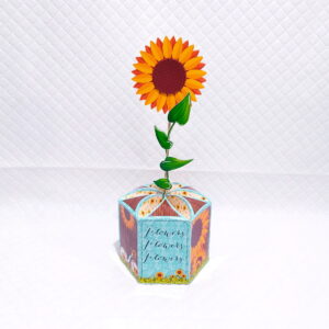 Girassol de papel em vaso hexagonal decorado, sobre fundo branco quadriculado.