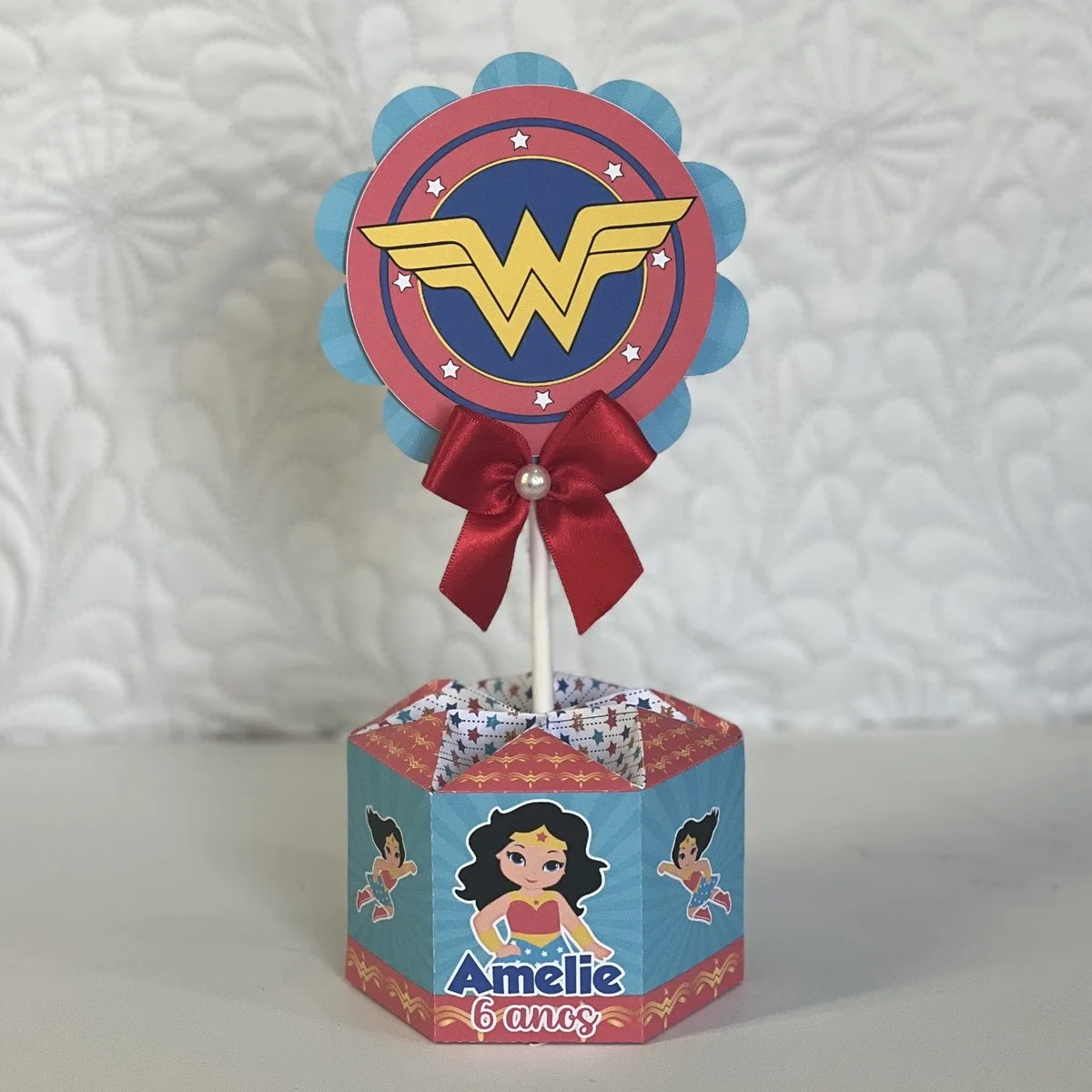 Decoração de mesa personalizada com tema de super-heroína e laço vermelho sobre base.