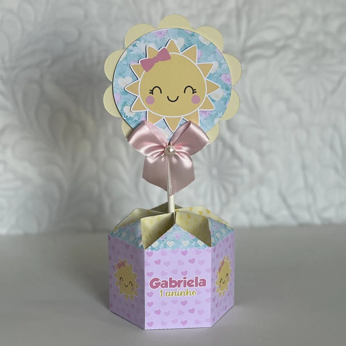 Centro de Mesa Sunshine Menina 1 Centro de mesa hexagonal com desenho de sol sorridente e laço de fita rosa.