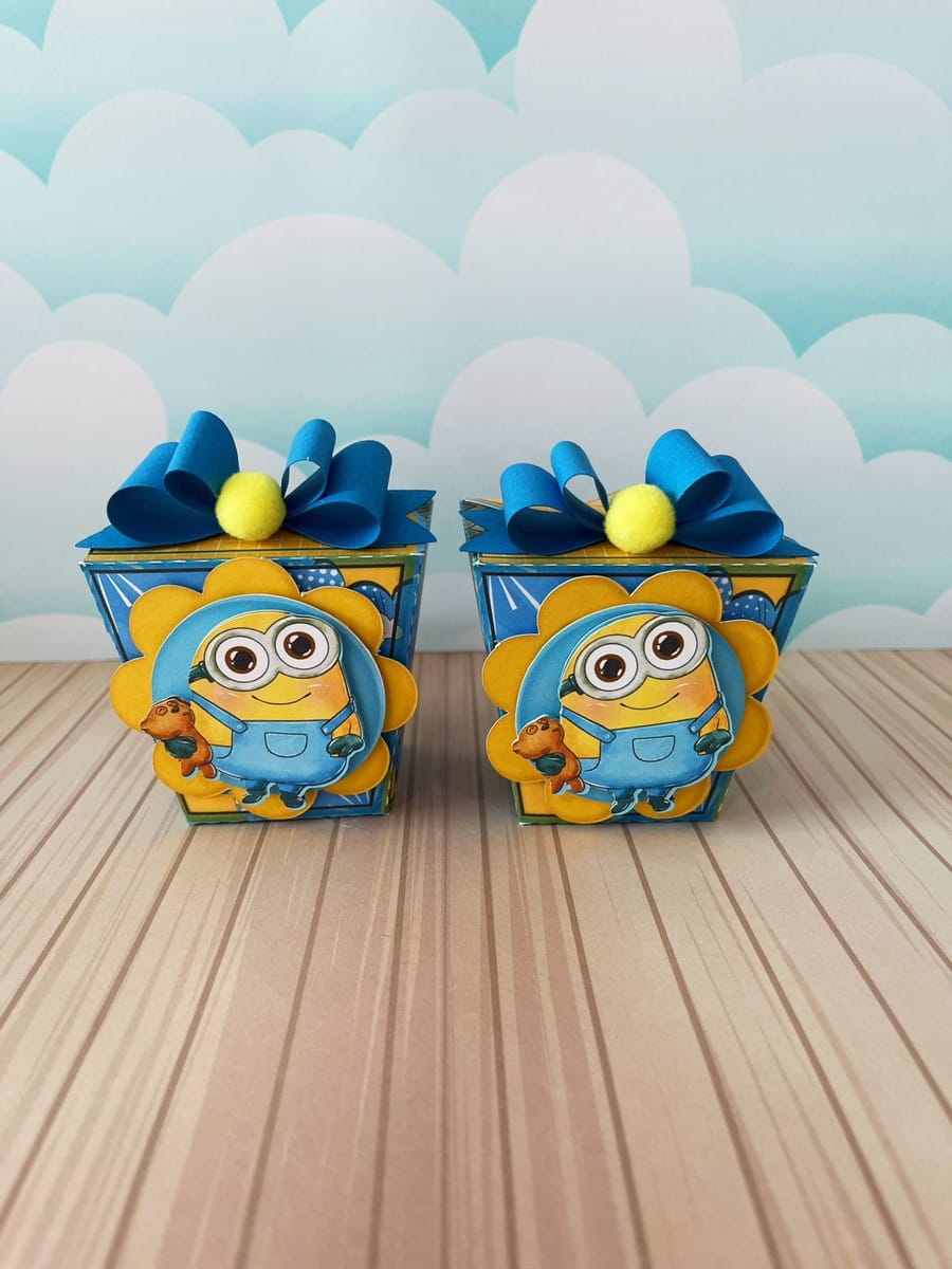 Kit Digital Festa Minions Luxo para Corte e Impressão 3 Duas caixas personalizadas decoradas com personagens amarelos, laços azuis e pompons amarelos sobre mesa.