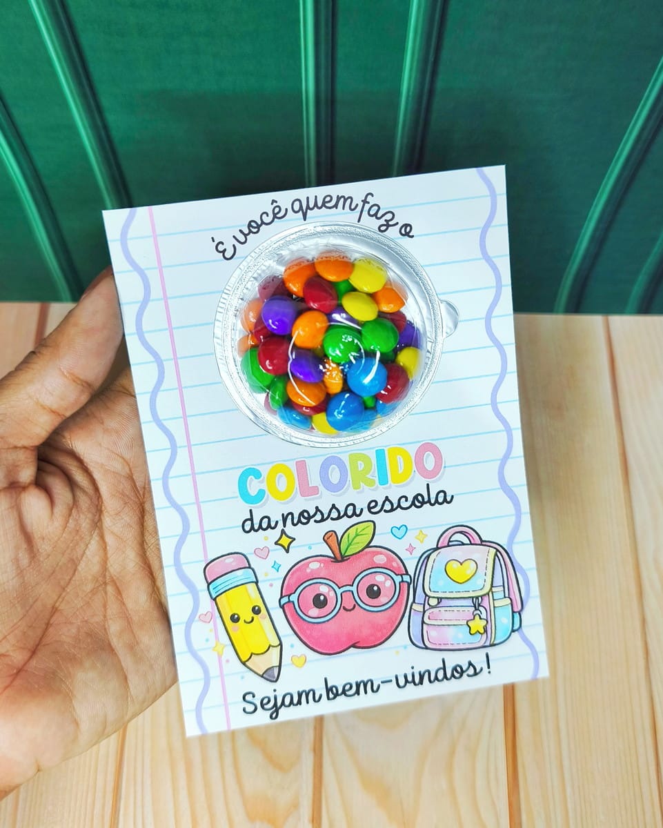Kit Digital Mimos Escolares Volta às Aulas 2026 4 Mão segurando cartão colorido de boas-vindas com guloseimas e ilustrações escolares.