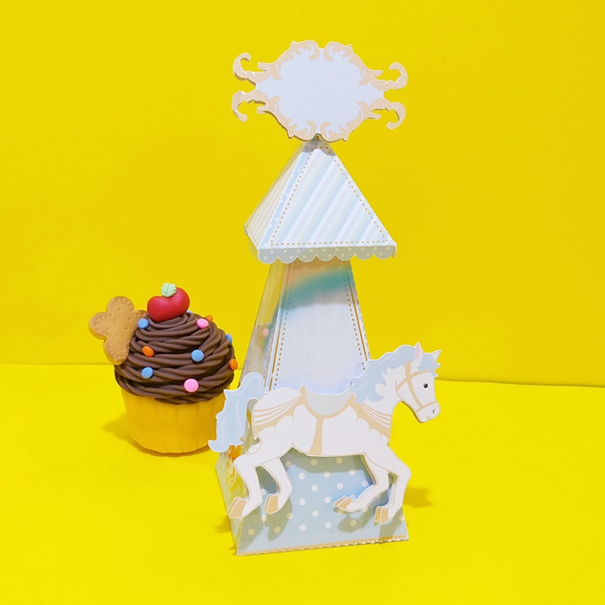 Kit Festa Carrossel Digital com Arquivos Editáveis 2 Uma torre de papel em formato cônico decorada com cavalo branco e um cupcake