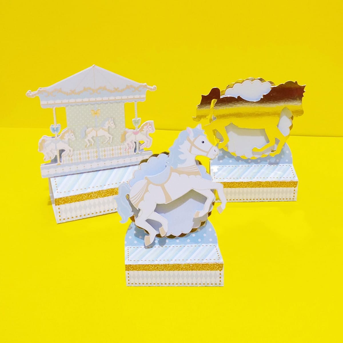 Kit Festa Carrossel Digital com Arquivos Editáveis 3 Três enfeites de papel com tema de carrossel em fundo amarelo.