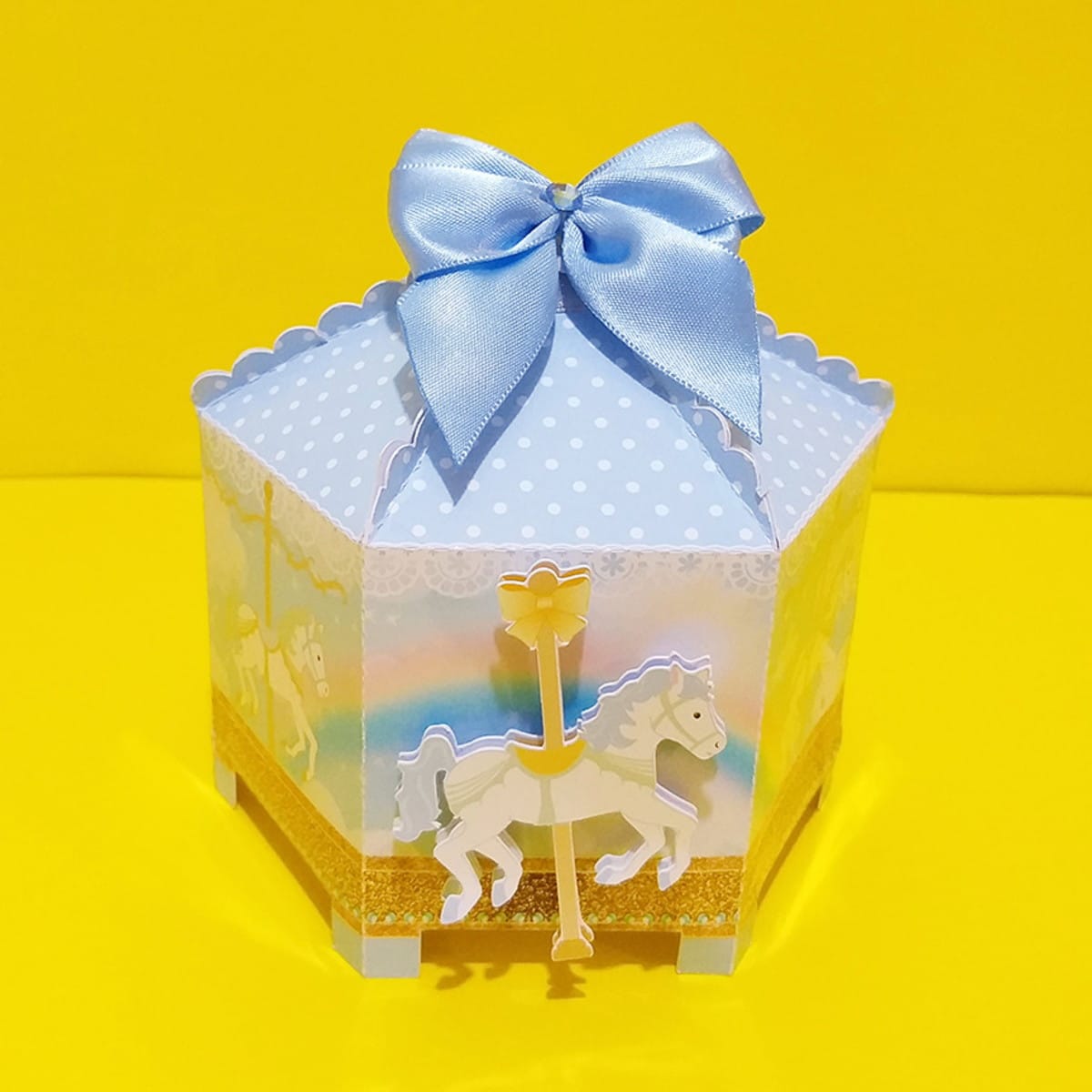 Kit Festa Carrossel Digital com Arquivos Editáveis 4 Caixa decorativa em formato de carrossel com laço azul e fundo amarelo.