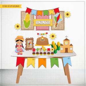 Decoração de festa temática com menina, milho, igreja, fogueira e bandeirinhas coloridas em mesa.