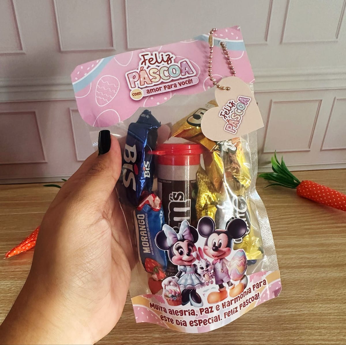 Kit Mimos de Páscoa Minnie e Mickey Digital 5 Mão segurando saquinho transparente com doces variados e tema de Páscoa infantil ilustrado.