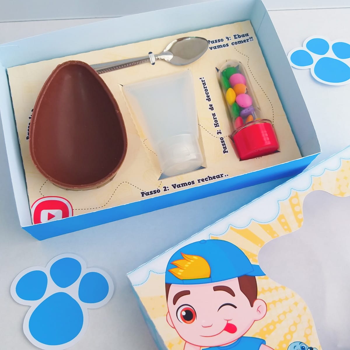 Kit Páscoa Luccas Neto Menino Arquivo Digital 2 Caixa azul contendo metade de um ovo de chocolate, colher, bisnagas e confeitos.