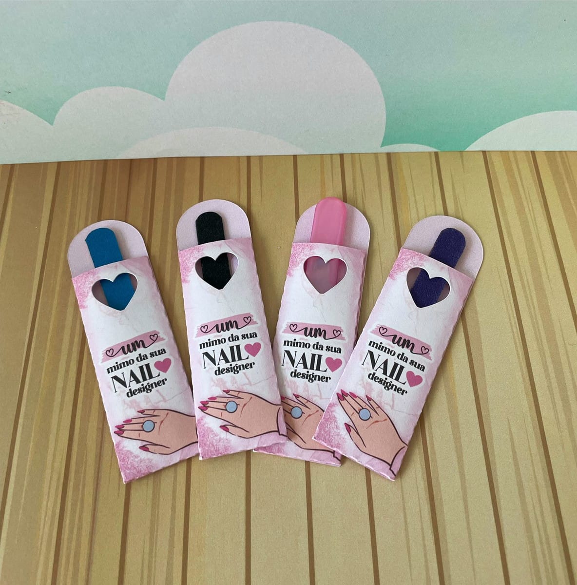 Mimos Nails Designer Arquivo Digital com Moldes de Corte 2 Quatro embalagens cor-de-rosa com lixas de unha coloridas dispostas sobre uma superfície amadeirada.