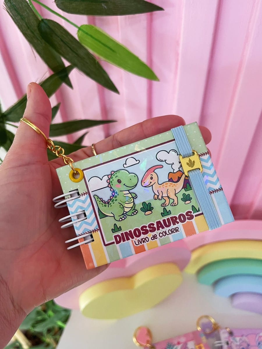 Mini Kit Dinossauro para Colorir Digital em PDF e PNG 2 Mão segurando um pequeno caderno de colorir com ilustração de dinossauros infantis.