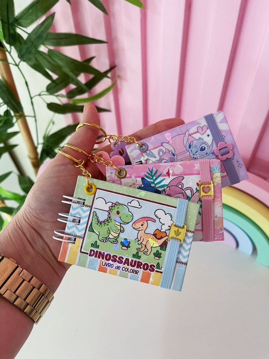 Mini Kit Dinossauro para Colorir Digital em PDF e PNG 3 Mão segurando pequenos cadernos coloridos com argolas metálicas em um fundo decorado