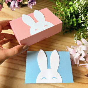 Mão segurando caixa de papel rosa e outra azul com coelhos recortados sobre madeira.