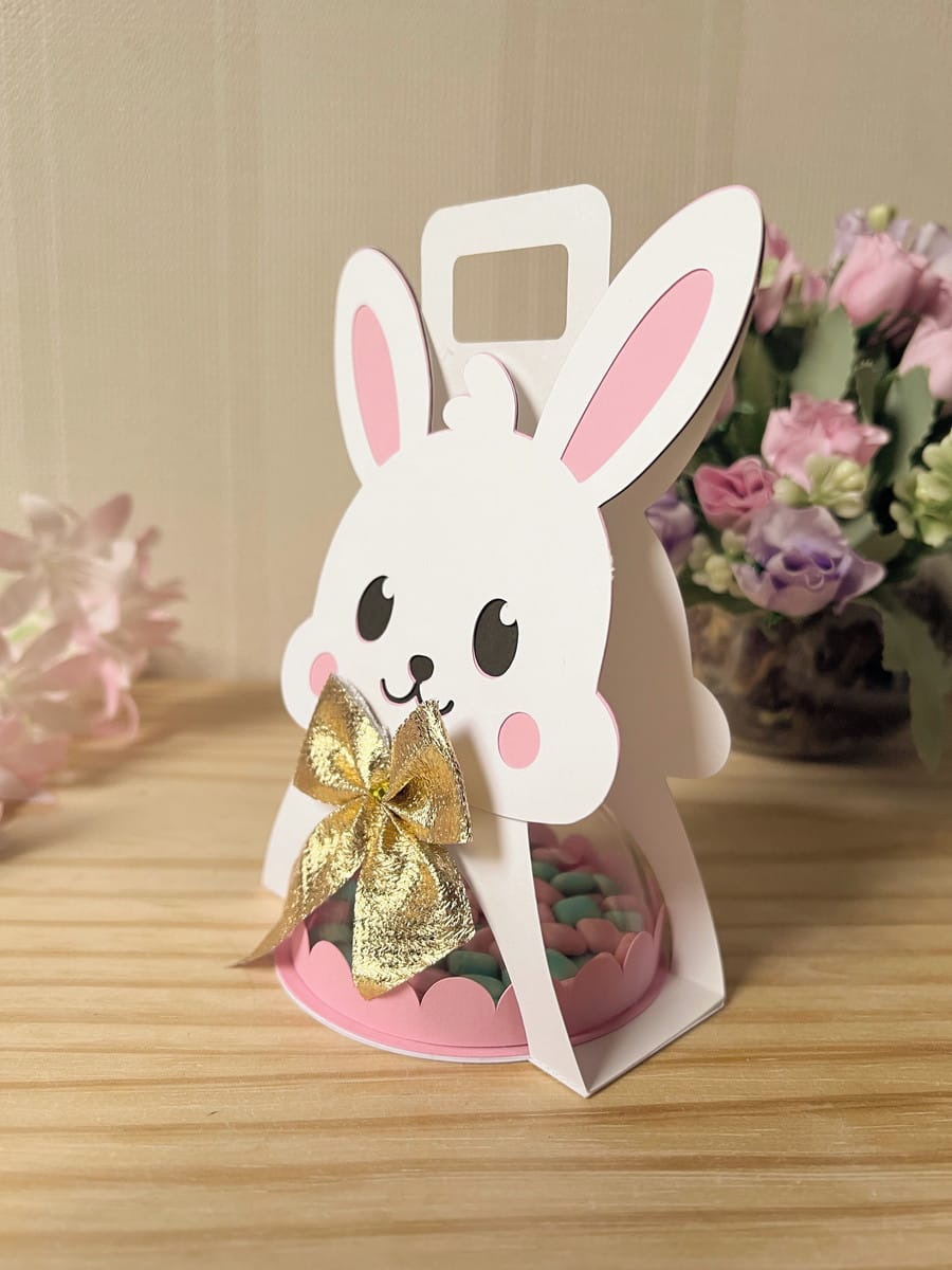 Sacolinha Coelhinha com Alça Arquivo Digital para Festas 2 Embalagem de papel em formato de coelho branco com laço dourado e doces coloridos.