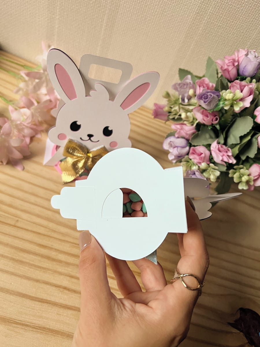 Sacolinha Coelhinha com Alça Arquivo Digital para Festas 3 Mão segurando uma embalagem de papel em formato de coelho com doces internos.