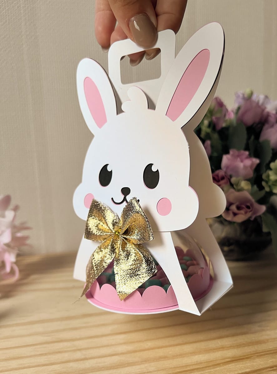 Sacolinha Coelhinha com Alça Arquivo Digital para Festas 5 Caixa de papel em formato de coelho branco com laço dourado sendo segurada.