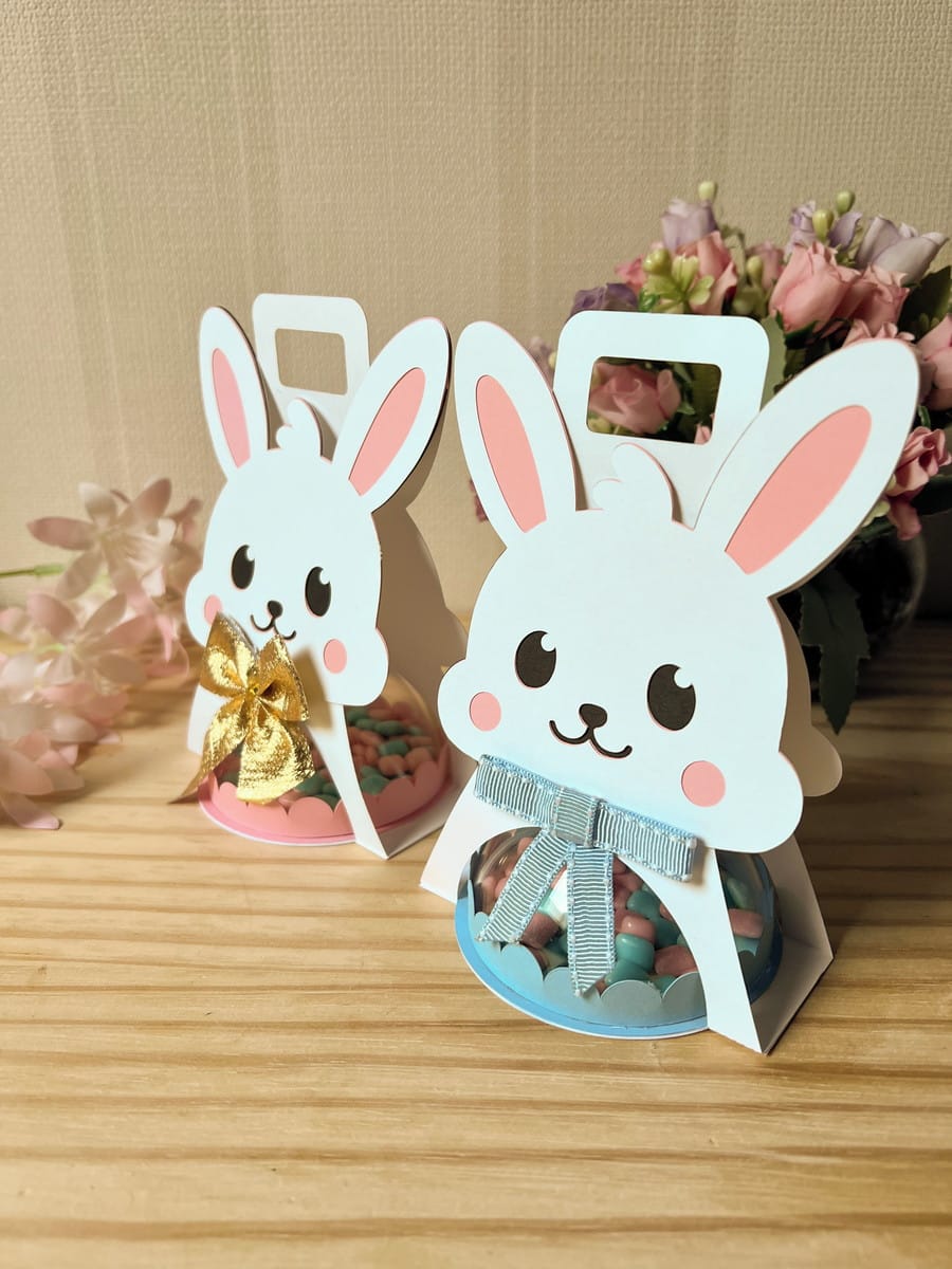 Sacolinha Coelhinha com Alça Arquivo Digital para Festas 1 Duas embalagens em formato de coelho contendo doces coloridos sobre uma mesa de madeira.