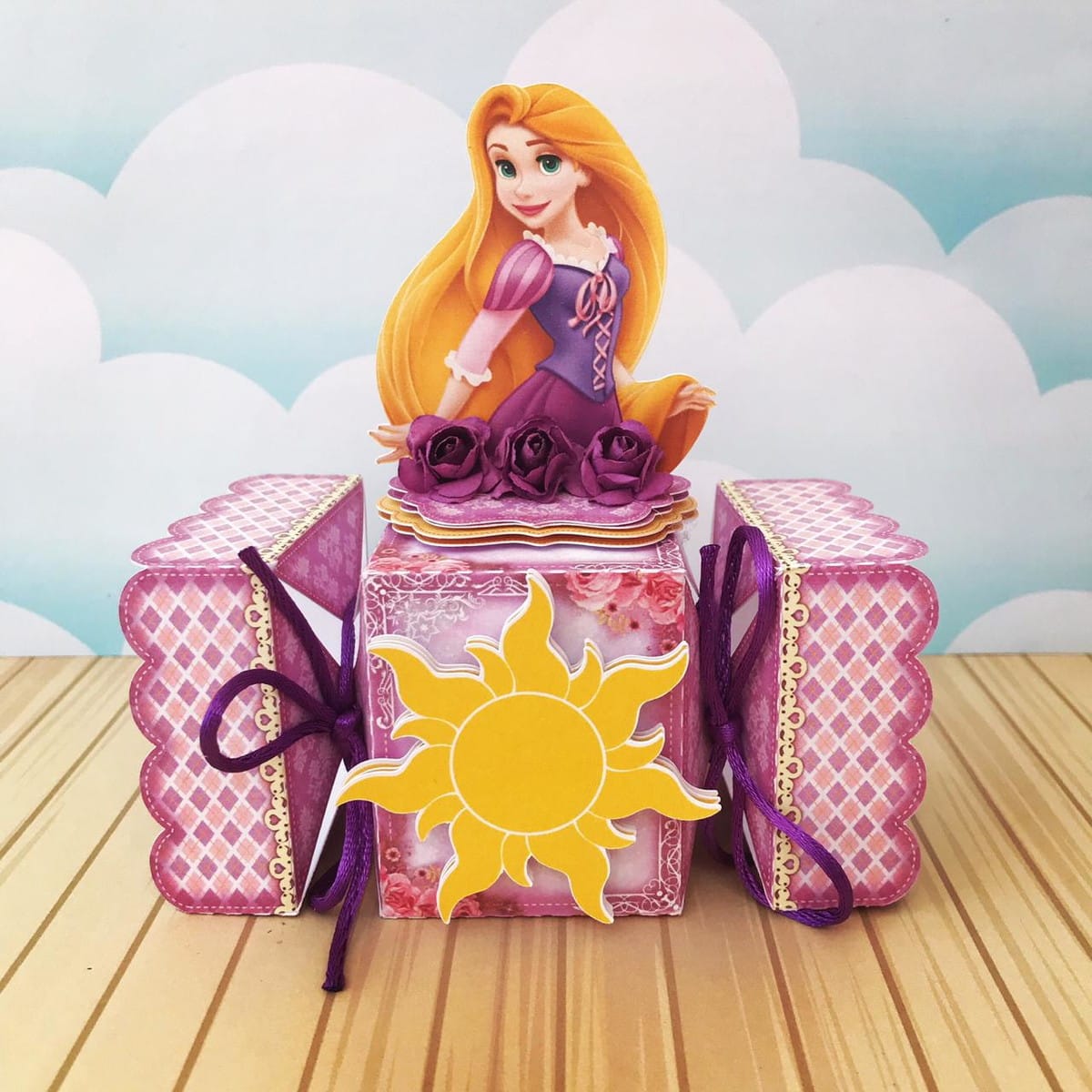 Arquivo Festa Rapunzel com Caixas Convite e Topo de Bolo 2 Caixa decorativa com personagem feminina de cabelo longo, flores roxas e sol amarelo.
